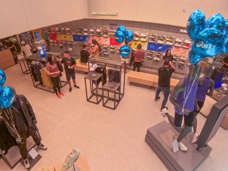 Imagem interna da loja Asics que exibe manequins segurando balões azul com o nome Asics escrito nos balões, em primeiro plano. No fundo da foto, há pessoas conversando e olhando produtos, além de uma prateleira com vários calçados.