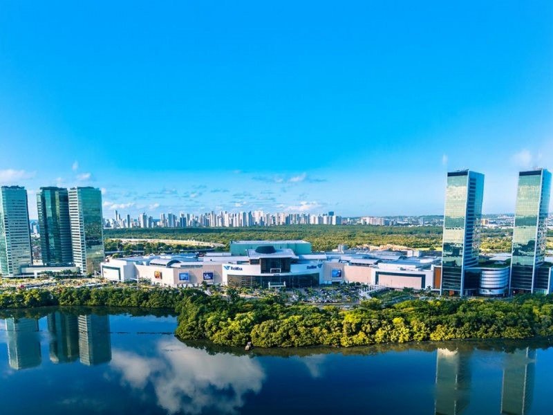 A imagem mostra uma foto do RioMar Recife a longa distância em um dia ensolarado com o céu azul. A foto também mostra as três torres empresariais do lado esquerdo e as outras duas torres do lado direito.