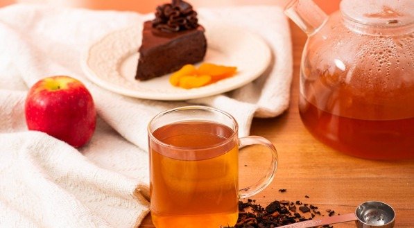 Imagem mostra um bule e uma xícara de chá, uma maçã e uma fatia de torta de chocolate com damascos ao lado