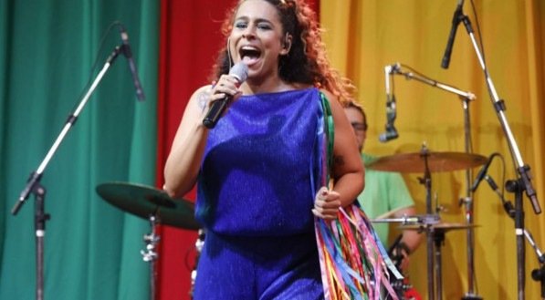 Uma mulher em um macacão azul canta no palco, segurando um microfone e fitas coloridas. Atrás dela, há uma bateria e cortinas verdes, vermelhas e amarelas.


