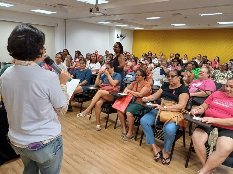 Uma mulher fala em um microfone enquanto encara um grande grupo de mulheres sentadas em uma sala de aula. Muitas participantes estão fazendo anotações e ouvindo atentamente. A sala tem piso de madeira e uma parede amarela de destaque.