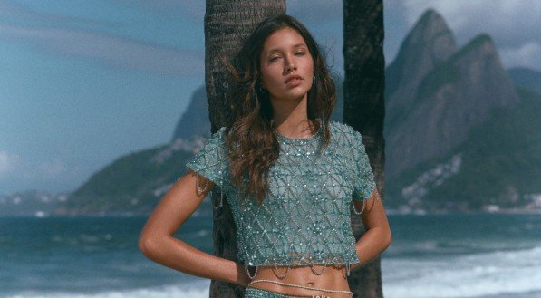 Uma modelo com top cropped e minissaia turquesa cravejados de mi&ccedil;angas posa na praia, com coqueiros e o Morro Dois Irm&atilde;os ao fundo.