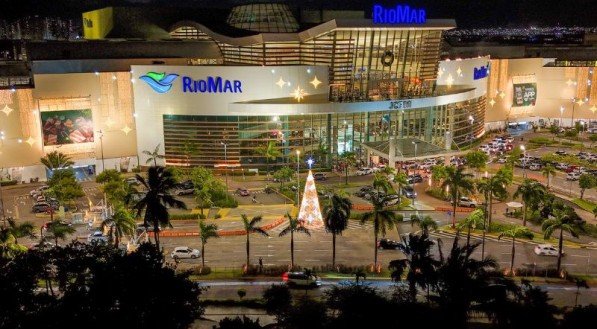 Vista noturna do RioMar Recife com fachada iluminada, carros estacionados, palmeiras e uma árvore de Natal iluminada em frente.