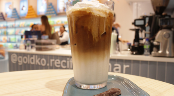 Copo com bebida gelada com café em um prato com um chocolate ao lado. O prato está em cima de uma mesa que tem a loja GoldKo de fundo.