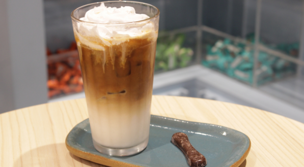 Bebida Iced Vanilla Latte em copo apoiado em cima de um prato com um chocolate GoldKo na lateral. Tudo está em uma mesa com chocolates GoldKo de fundo.