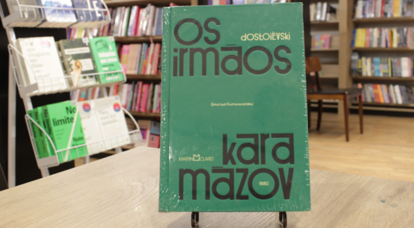 Livro "Irmãos Karamazov", do autor Fiódor Dostoiévski, em uma mesa com livros em estante ao fundo.