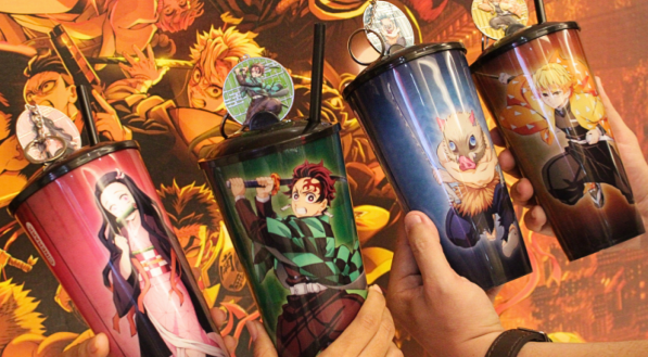 Quatro mãos seguram copos coloridos com desenhos de personagens do anime Demon Slayer. Cada copo apresenta um personagem diferente. O fundo mostra um pôster vibrante do anime.