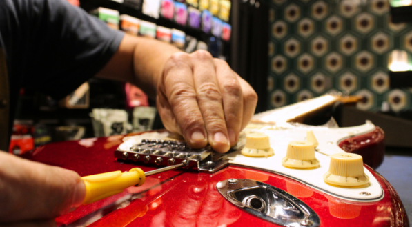M&atilde;os sobre a guitarra com uma ferramenta realizando conserto no instrumento com um fundo desfocado.
