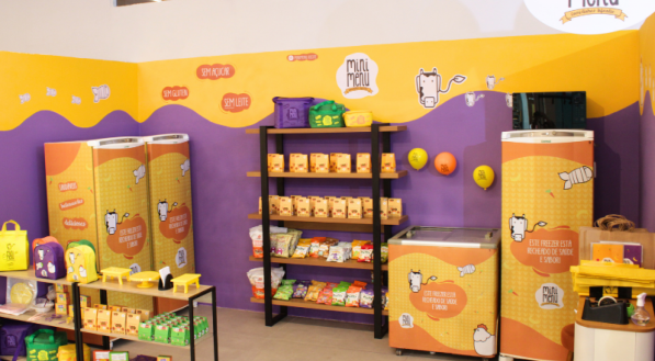 Um canto de loja com cores vivas, com paredes amarelas e roxas, exibindo a marca "Mini Menu". As prateleiras contêm lanches, coolers, mochilas e produtos com personagens de desenhos animados e designs divertidos.