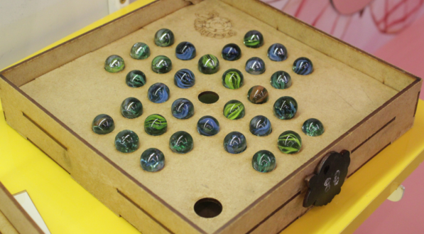 Jogo resta um, feito em madeiro e com bolas de gude, em uma mesa amarela.