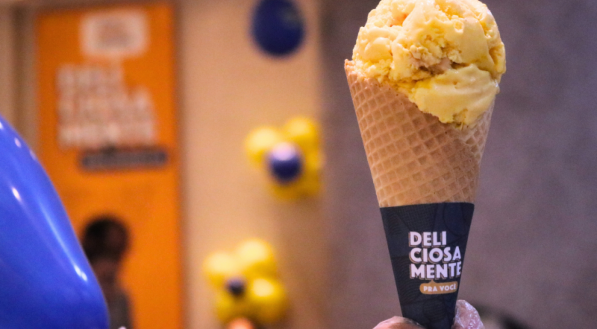 Casquinha de sorvete com uma bola de sorvete na cor marrom clara, no sabor de doce de leite, e uma colher pequena na cor branca na bola de sorvete