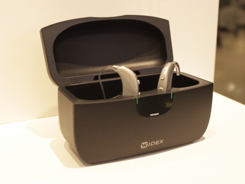 A imagem mostra dois aparelhos auditivos da marca Widex posicionados dentro de um estojo carregador preto com a tampa aberta.