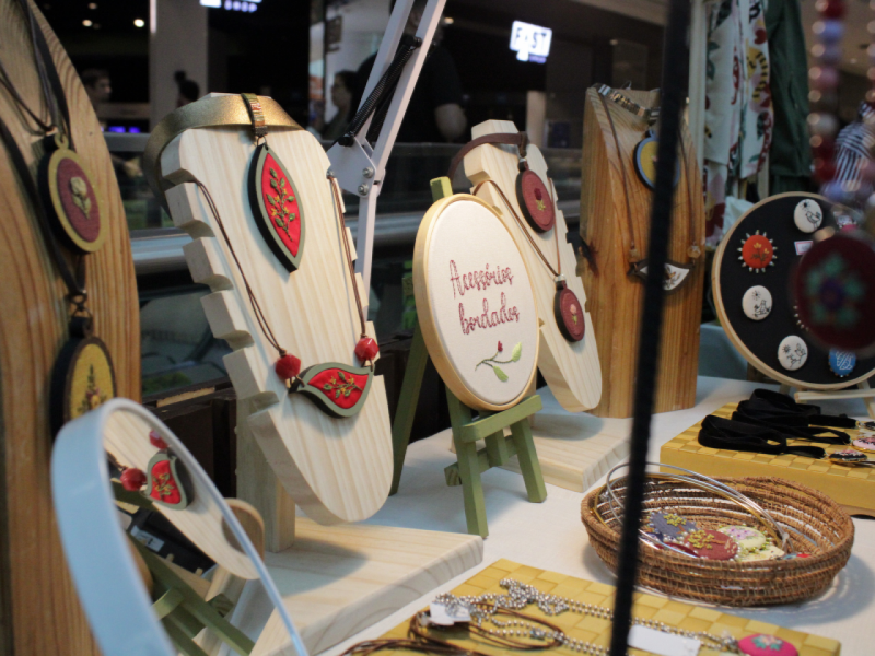 A imagem mostra um expositor de joias e colares artesanais de madeira e bordados. 
