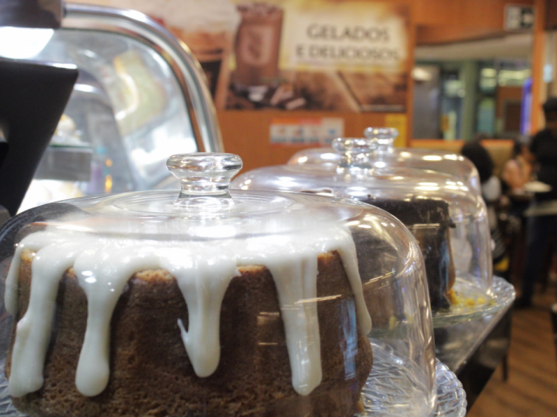 A imagem mostra vários bolos com coberturas escorrendo são exibidos sob cúpulas de vidro em um balcão.