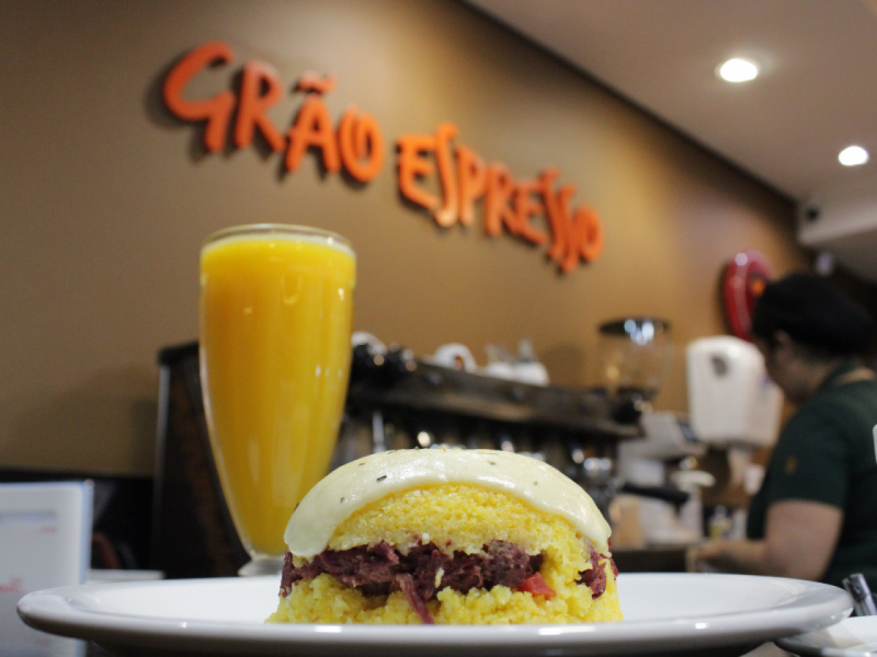 A imagem mostra um cuscuz recheado com charque e coberto com queijo em primeiro plano. Ao fundo, é possível ver o letreiro 