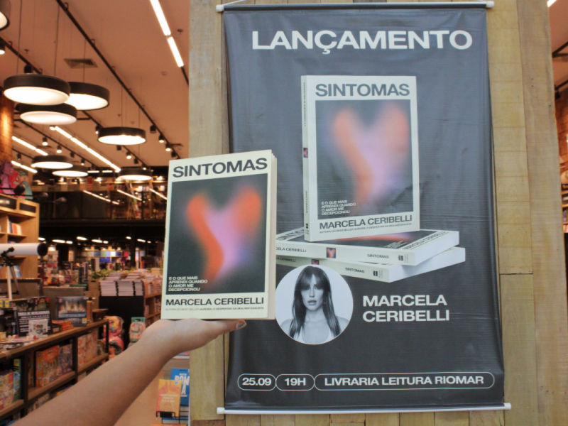  Um exemplar do livro "Sintomas" é segurado em frente a um banner do lançamento na Livraria Leitura do RioMar Recife.