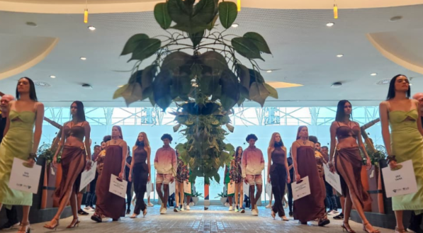 Modelos desfilam em um sal&atilde;o. Eles usam roupas leves em tons de verde, marrom e lil&aacute;s, com estampas de gradiente.