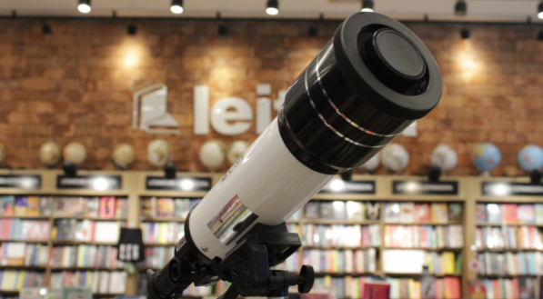 Um telescópio branco e preto com tripé, exposto sobre livros em uma livraria.