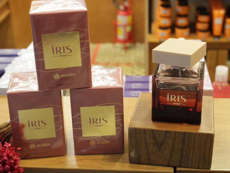 A imagem mostra o perfume "Íris Rose" da Avatim, ao lado de três embalagens do mesmo fechadas sobre um balcão de madeira.