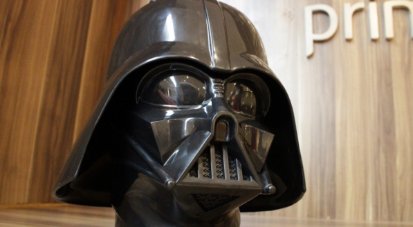 Uma foto frontal e próxima do balde de pipoca em formato de capacete do Darth Vader. O balde está sobre uma superfície e o fundo é uma parede de madeira com a palavra "bradesco" visível.