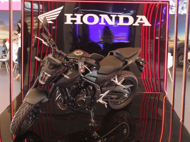 A imagem mostra uma moto Honda preta em um estande com iluminação de LED vermelha e um fundo espelhado.
