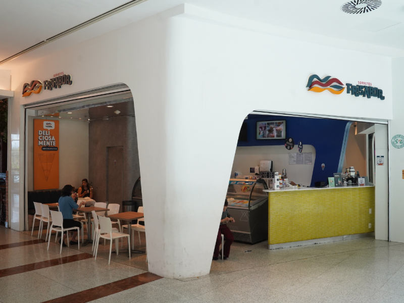 A imagem mostra a fachada da FriSabor, com uma entrada em formato de "L" e um balcão de atendimento amarelo. 