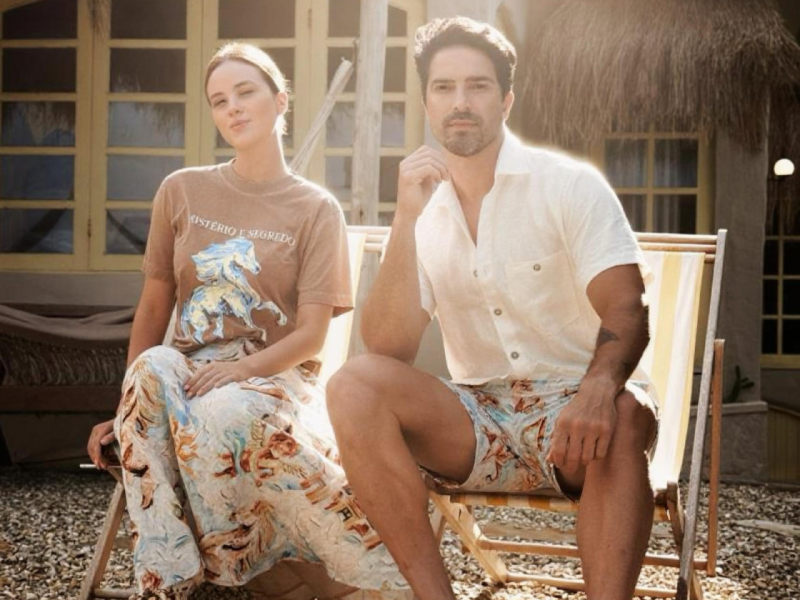 A imagem mostra um casal, sentados em cadeiras de praia, usando peças da coleção 'Pelas Ruas que Andei'.