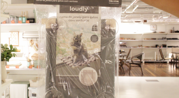 Uma mão segura uma embalagem plástica de cama de janela para gatos com ventosas, da marca Loudly Tok&Stok. A embalagem mostra a imagem de um gato deitado na cama.