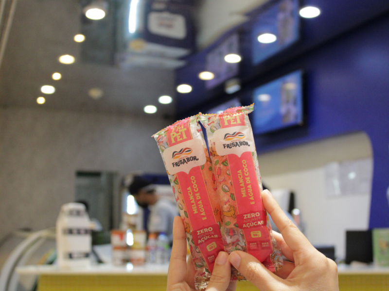 Duas mãos seguram dois picolés para pet da marca Frisabor, sabor melancia e água de coco, com embalagem rosa. O fundo é de uma loja com iluminação embutida.