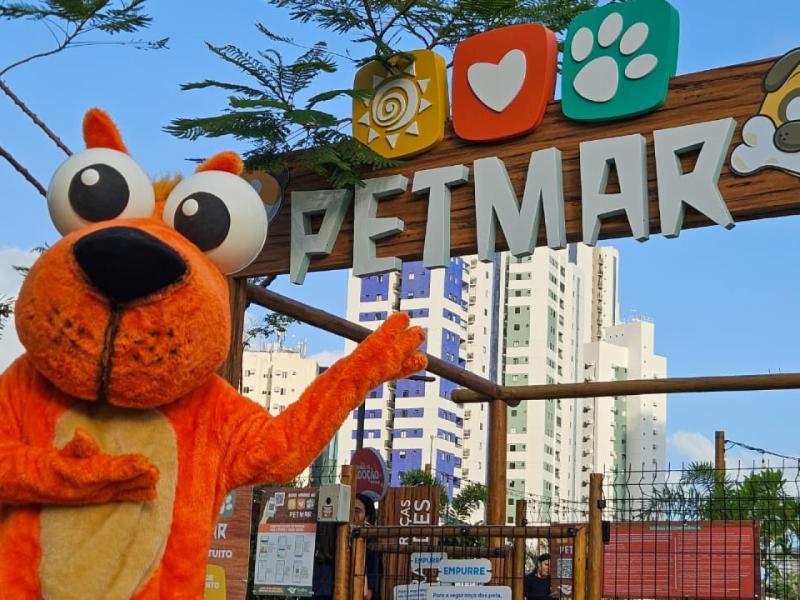  O mascote laranja sorridente em frente à placa de entrada do evento "PETMAR", com prédios da cidade ao fundo.