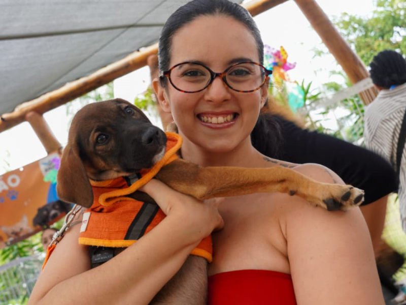 Tayane sorrindo, de óculos, abraçando carinhosamente a filhote Maria Flor, que está usando um colete laranja.