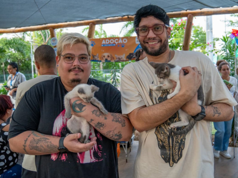 Esli e Carlos sorridentes segurando duas filhotes de gato no colo, em uma área coberta do evento de adoção.