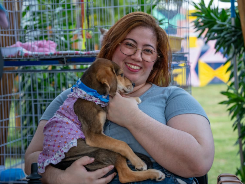 Maria Helena sorridente abraçando uma filhote de cachorro que usa uma roupinha xadrez.