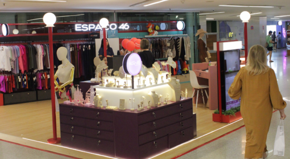 Vista angular de um quiosque de moda e acessórios iluminado. Há araras de roupas no fundo, um balcão central com joias e uma pessoa de vestido mostarda caminhando à direita.