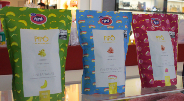 Três pacotes de pipoca Pipó Fini estão lado a lado em um balcão: bananas (verde), dentaduras (azul) e beijos (rosa).