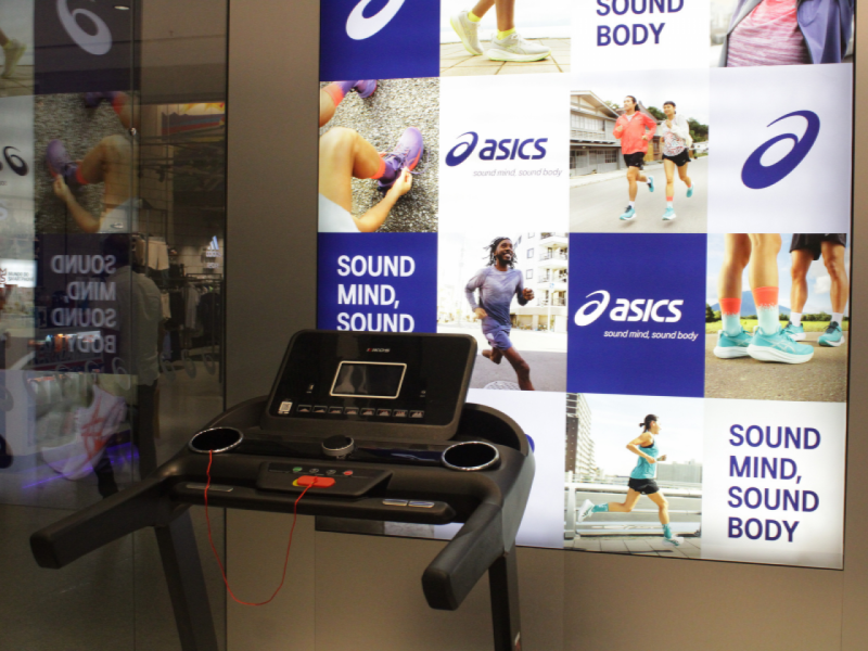 Uma esteira de exercícios está posicionada em frente a um grande mural da Asics.