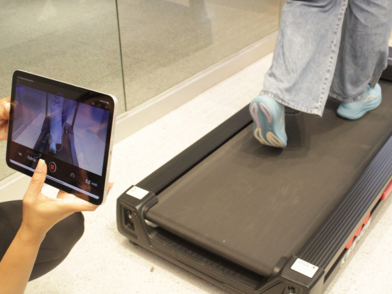 Uma pessoa usando tênis azuis caminha sobre uma esteira enquanto é feita uma análise do caminhar através de um tablet