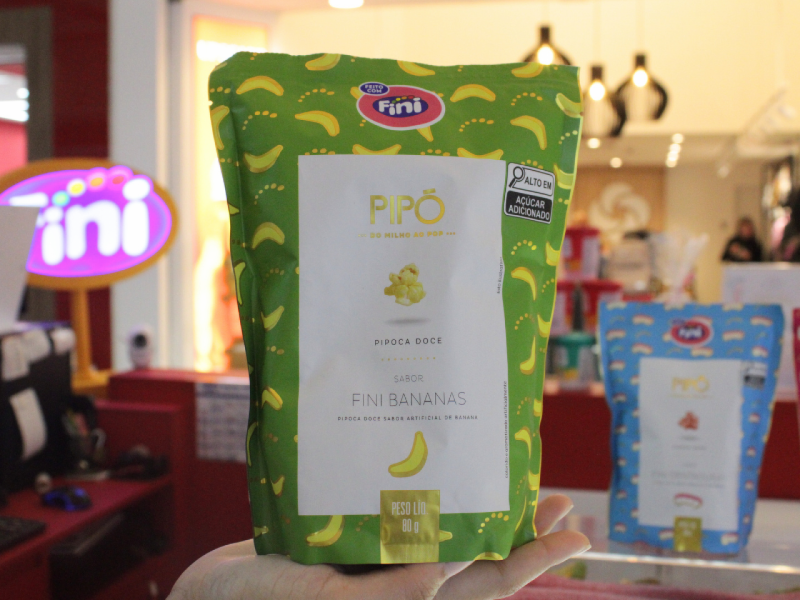 Uma mão segura em destaque um pacote de Pipó Fini Bananas (pipoca doce com sabor de banana), com embalagem verde.
