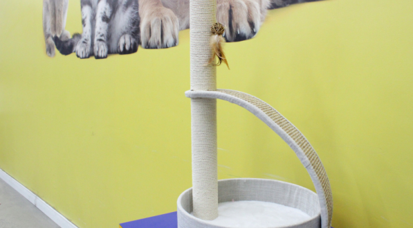 A imagem mostra um arranhador multifuncional com um poste de sisal, um brinquedo de pena e uma base que serve como caminha acolchoada.