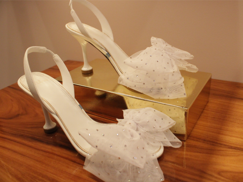 A imagem mostra um par de sandálias slingback brancas de salto fino, adornadas com grandes laços de tule pontilhados de brilho.