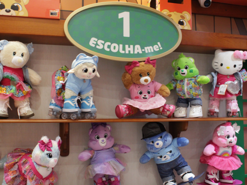 A imagem mostra a etapa 1 (escolha-me!) e as prateleiras com diversas pelúcias, o primeiro passo para selecionar o modelo base da sua criação.