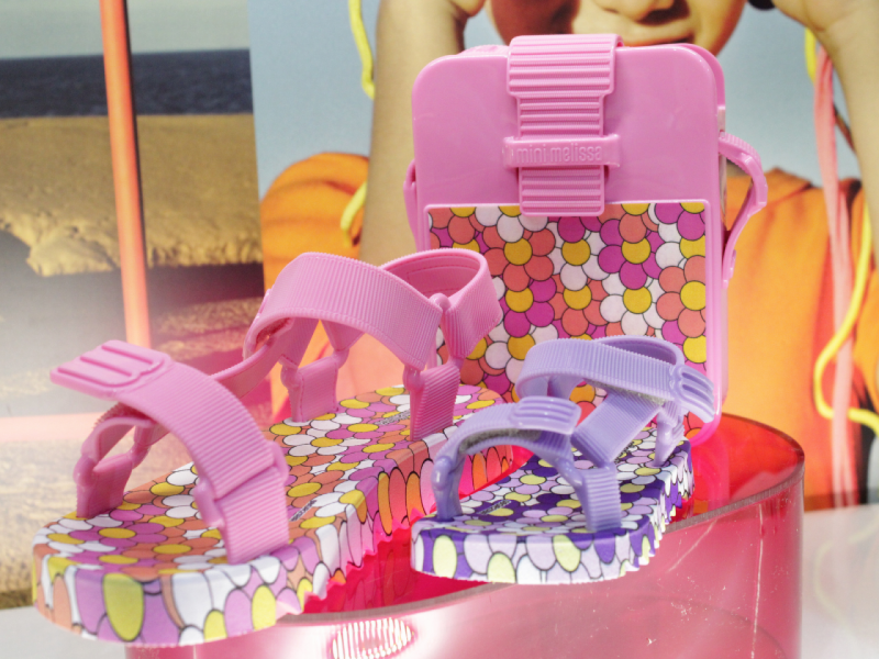 A imagem mostra duas sandálias, nas cores rosa e roxo com estampas de bolinha, posicionadas ao lado de uma mini-bolsa tiracolo rosa da mesma estampa.