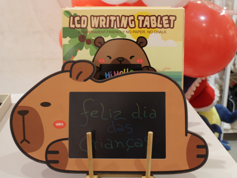 A imagem mostra uma lousa digital em formato de capivara exibe a frase "Feliz dia das crianças" ao lado da embalagem do produto.