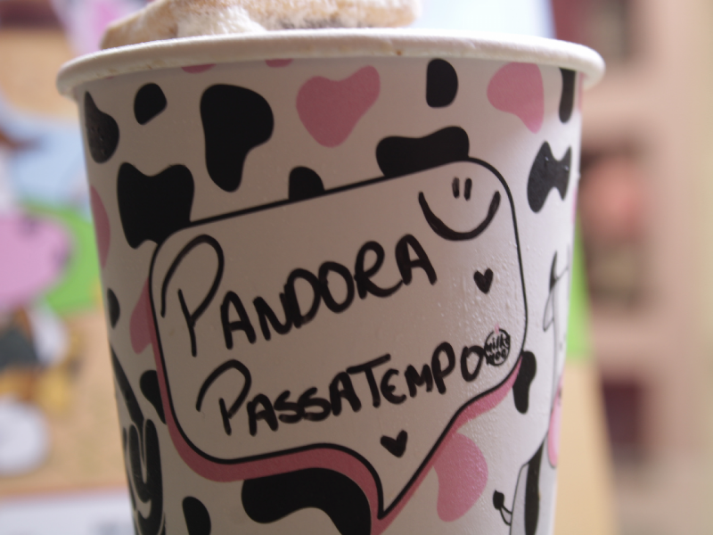 Foto de um copo de milkshake com estampa de vaquinha com "Pandora Passatempo" escrito. A bebida tem um biscoito no topo.