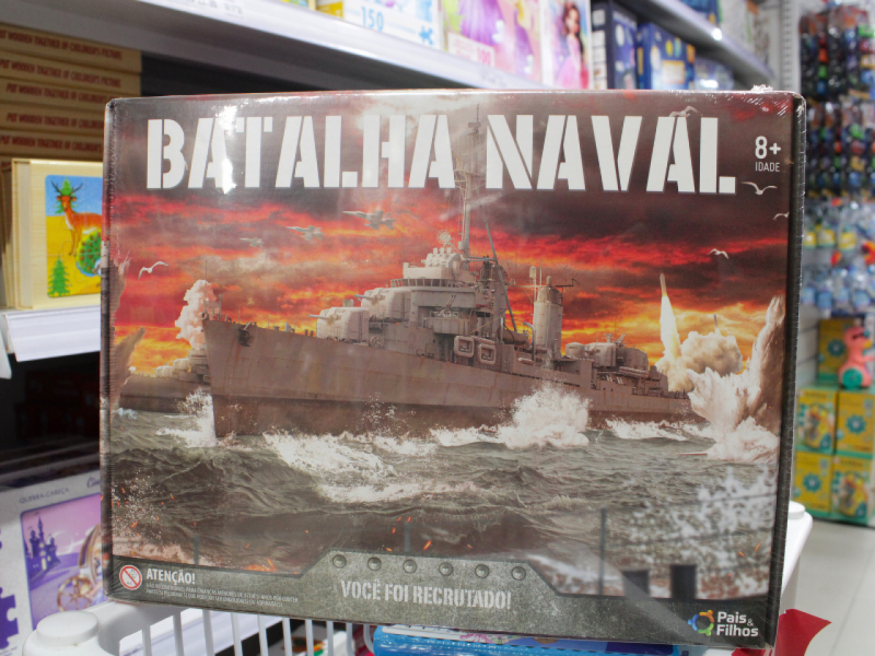 A imagem mostra a caixa do jogo de tabuleiro "Batalha Naval", com uma ilustração  de um navio de guerra em combate ao pôr do sol.