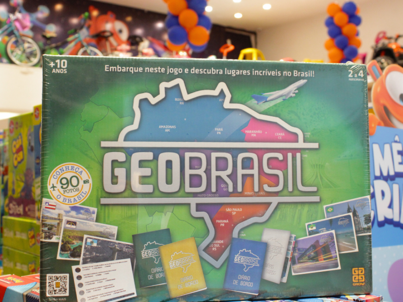 A imagem mostra a caixa do jogo de tabuleiro "GeoBrasil" da Grow, com o contorno do mapa do Brasil e elementos de viagem como aviões e postais.