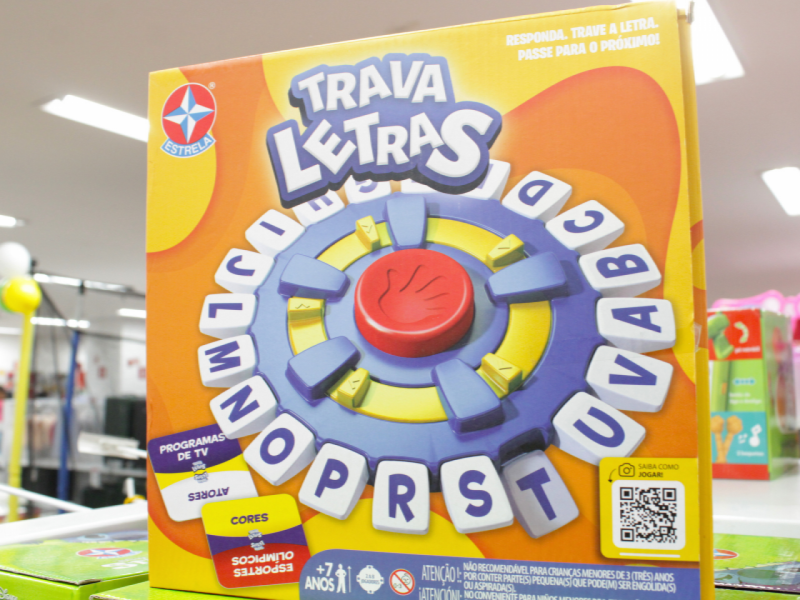 A imagem mostra a caixa do jogo "Trava Letras" da Estrela, com um design circular colorido representando as letras móveis do jogo de palavras.