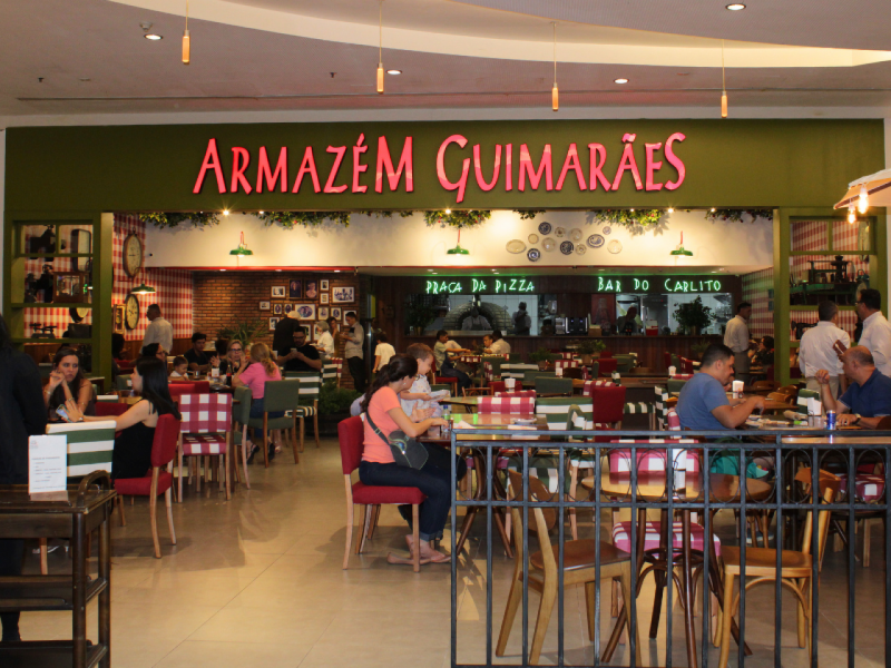 A imagem mostra a fachada interna e a área de refeições do restaurante "Armazém Guimarães" no RioMar Recife , com mesas e cadeiras de cores vivas e xadrez.