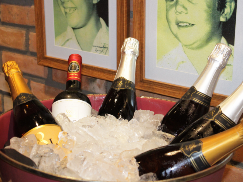 A imagem mostra uma seleção de garrafas de vinho, incluindo espumantes e um vinho tinto, resfriando em um balde vermelho cheio de gelo.