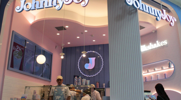 A imagem mostra a fachada da loja Johnny Joy com a logomarca iluminada, cores pastel e funcionários atendendo clientes no balcão.